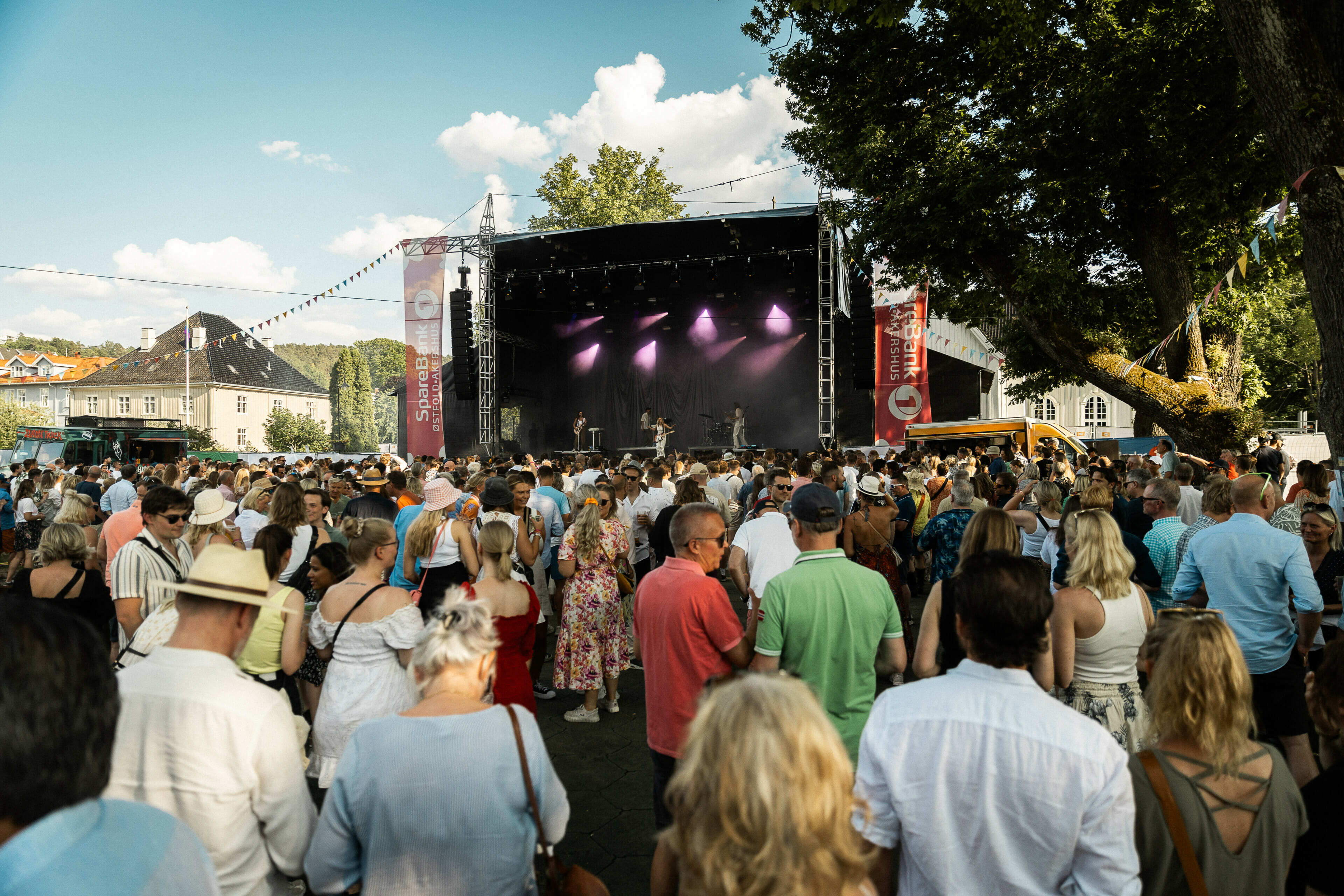 Bildet viser en dagskonsert under Drøbakfestivalen 2023. Bildet er tatt slik at man ser hovedscenen og noen hundre publikummere som følger med på konserten. Det er dagtid og sola skinner.