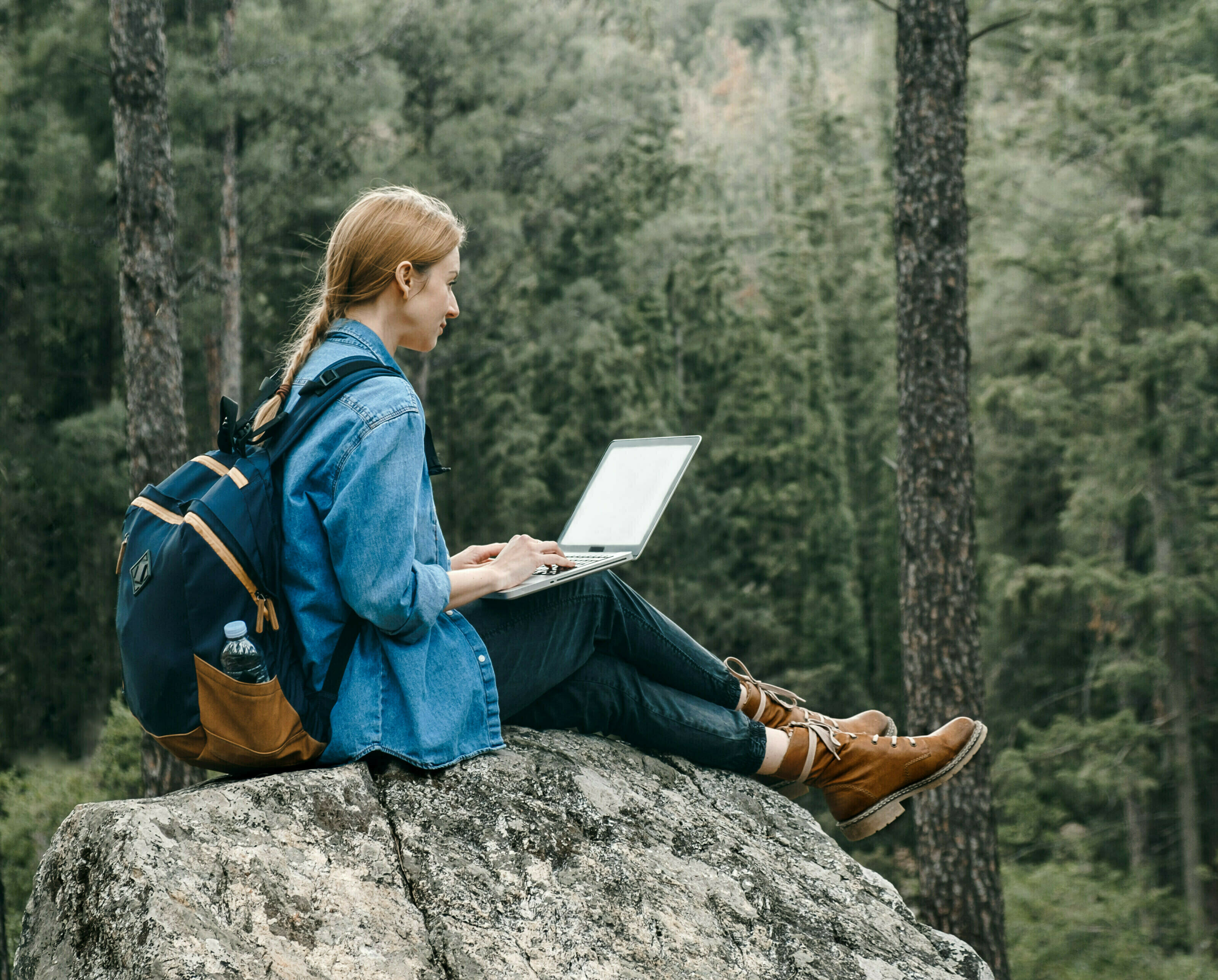 Young woman with backpack sitting on ground at forest and working on laptop . Workation concept.