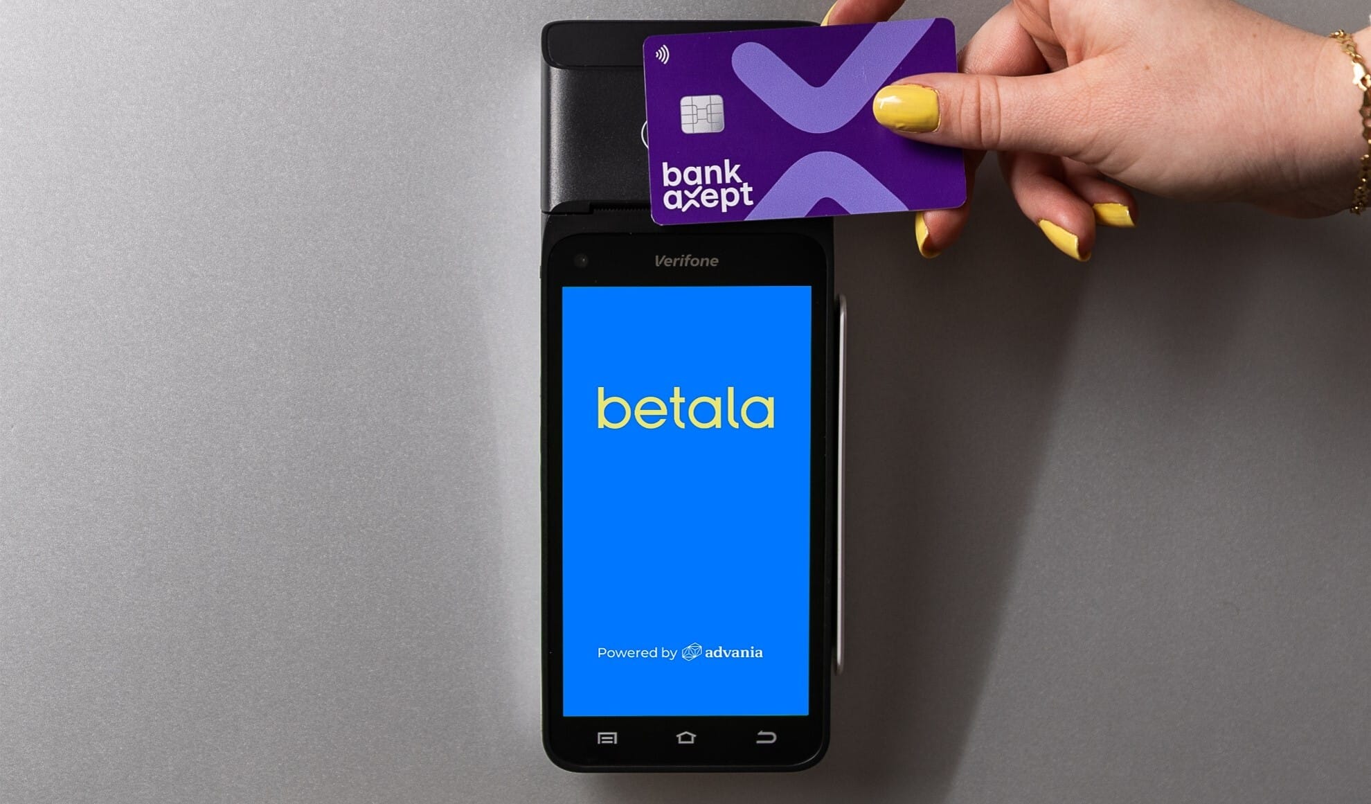 Bildet viser en betalingsterminal. På skjermen er Betala sin logo avbildet. Det er en hånd som "tæpper" et BankAxept betalingskort over betalingsterminalen.